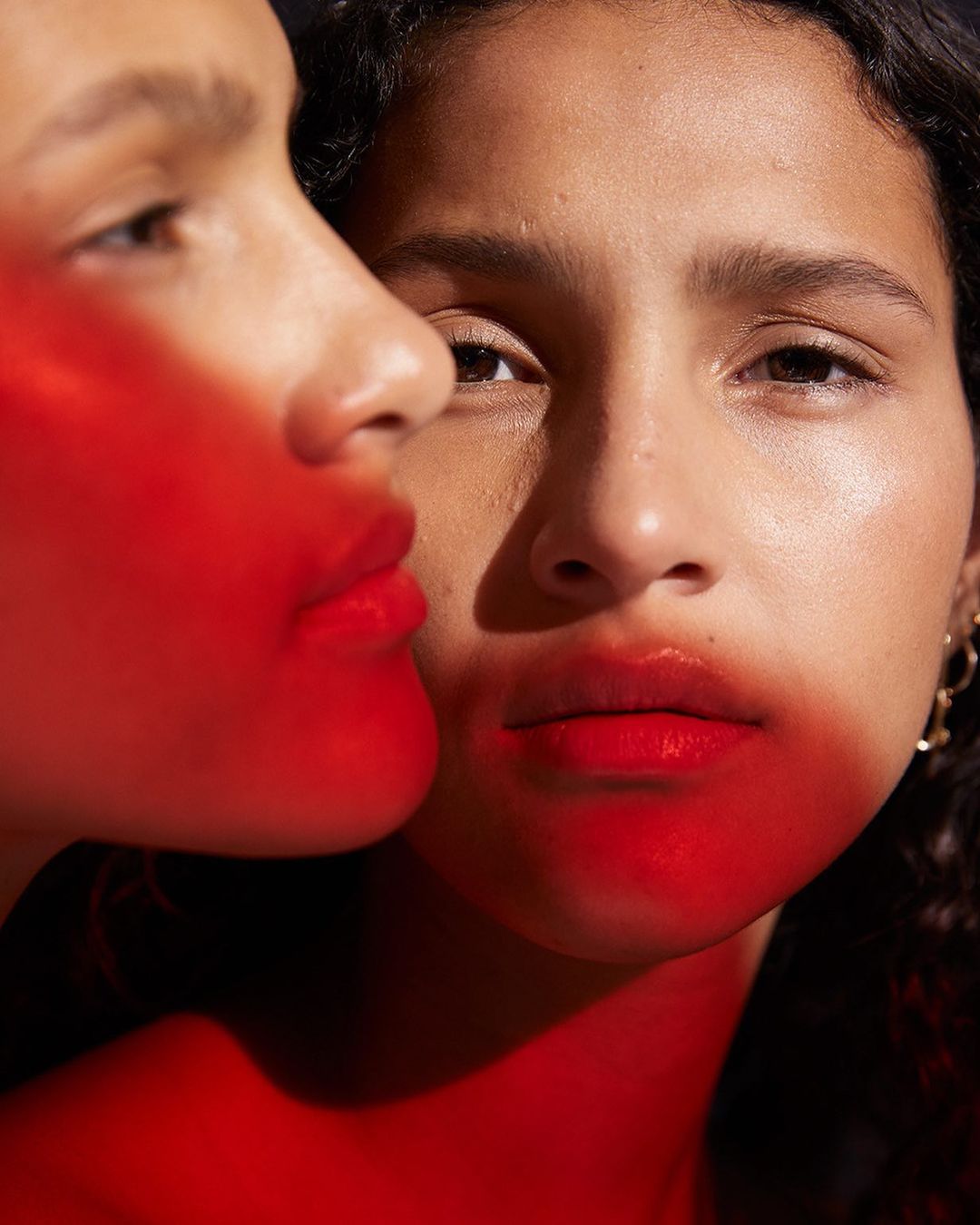 Close-up beauty shot featuring two young adult females with red lips and glowing skin in a dark studio, showcasing makeup and skincare.