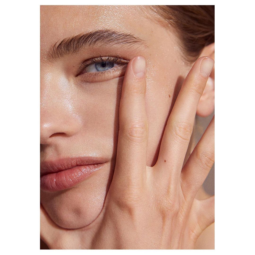 Close-up portrait of a fair-skinned adult female with blue eyes, featuring natural makeup and hand near face in soft, diffused lighting.