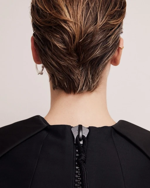 Back view of an adult female with short blond hair wearing a black top with a zipper; focus on fashion and hair details in a studio setting.