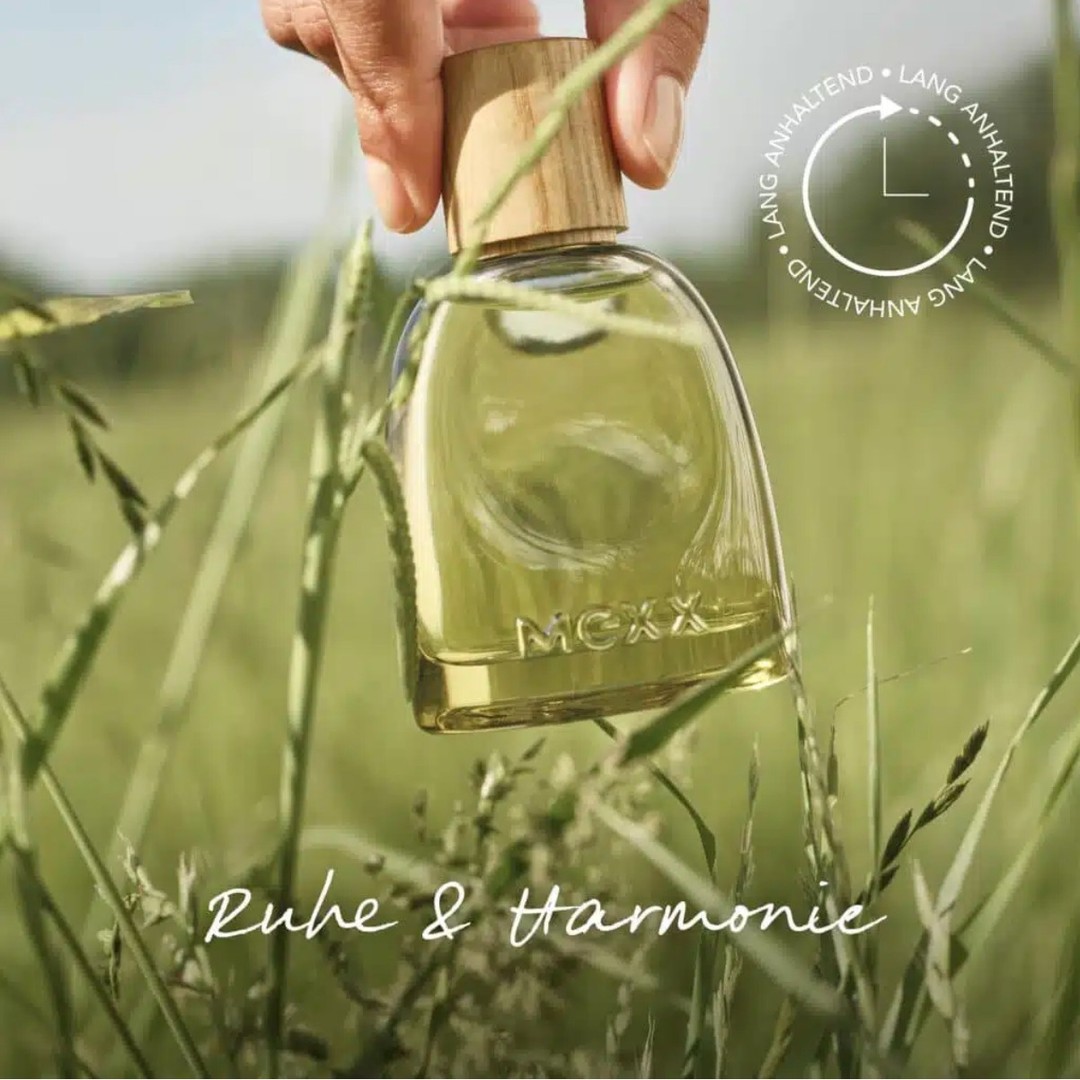 Close-up of a perfume bottle in a grassy field, held by a hand. Bottle has a wooden cap, with a blurred green field background.