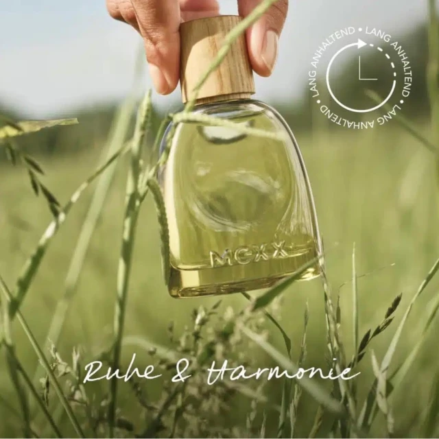 Close-up of a perfume bottle in a grassy field, held by a hand. Bottle has a wooden cap, with a blurred green field background.