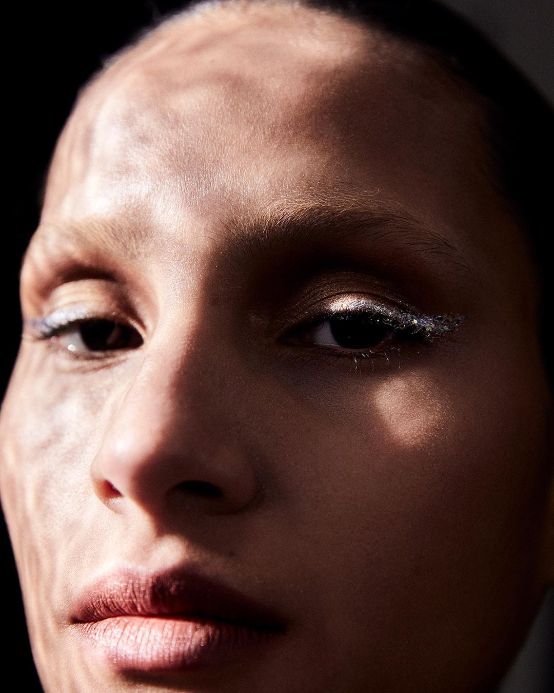 Close-up portrait of a woman's face with dramatic lighting, showcasing her features and makeup, set against a dark background.