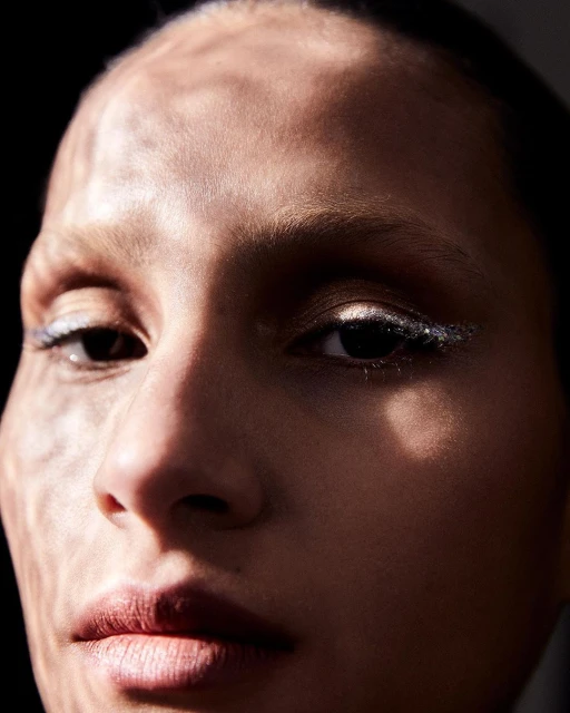 Close-up portrait of a woman's face with dramatic lighting, showcasing her features and makeup, set against a dark background.