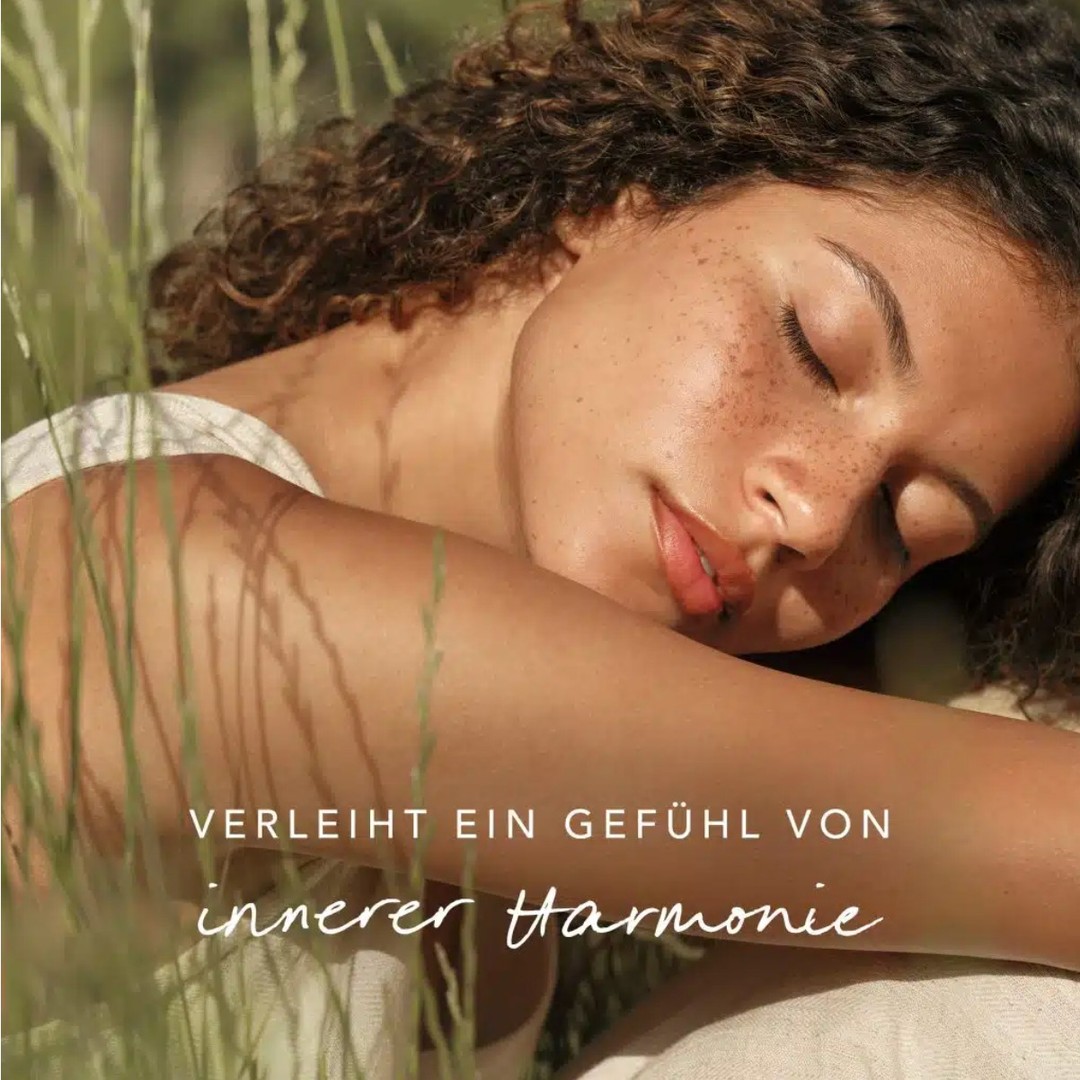A young adult woman with curly hair rests in tall grass in an outdoor setting, eyes closed, basking in the warm light.