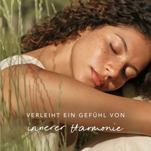 A young adult woman with curly hair rests in tall grass in an outdoor setting, eyes closed, basking in the warm light.