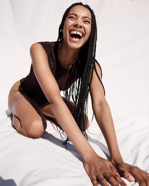 Portrait of a smiling young woman with dark braids wearing a dark top, kneeling on a white sheet in a bright, eye-level indoor shot.
