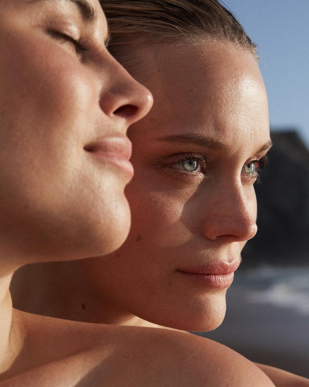 Close-up of two light-skinned women with natural makeup posing outdoors with soft light. One woman has her eyes closed. Beauty shot.
