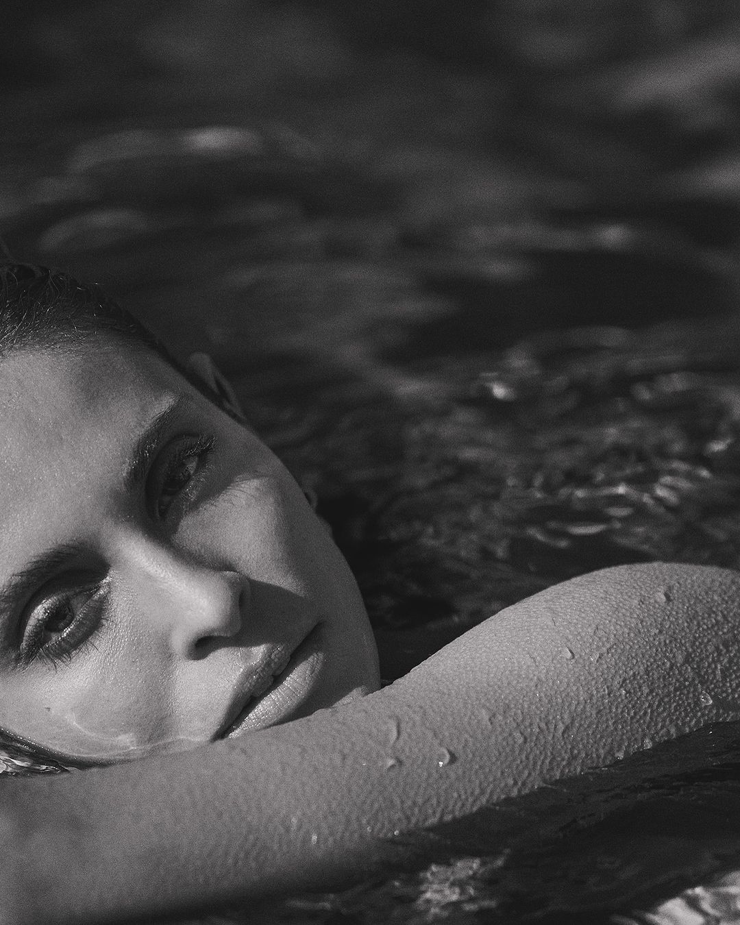 Close up monochrome portrait of a woman resting her arm on the water surface with water droplets visible on her skin.