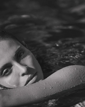 Close up monochrome portrait of a woman resting her arm on the water surface with water droplets visible on her skin.