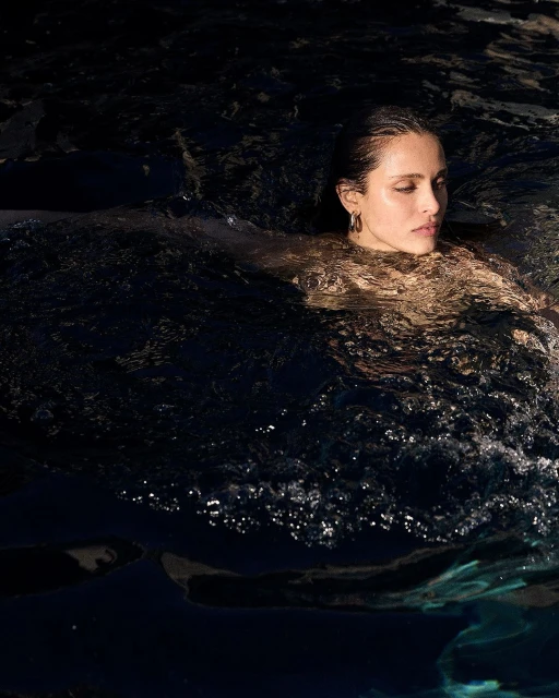 Adult female swims in dark water, looking away with calm expression, wearing dark earrings, in an indoor pool with natural lighting.