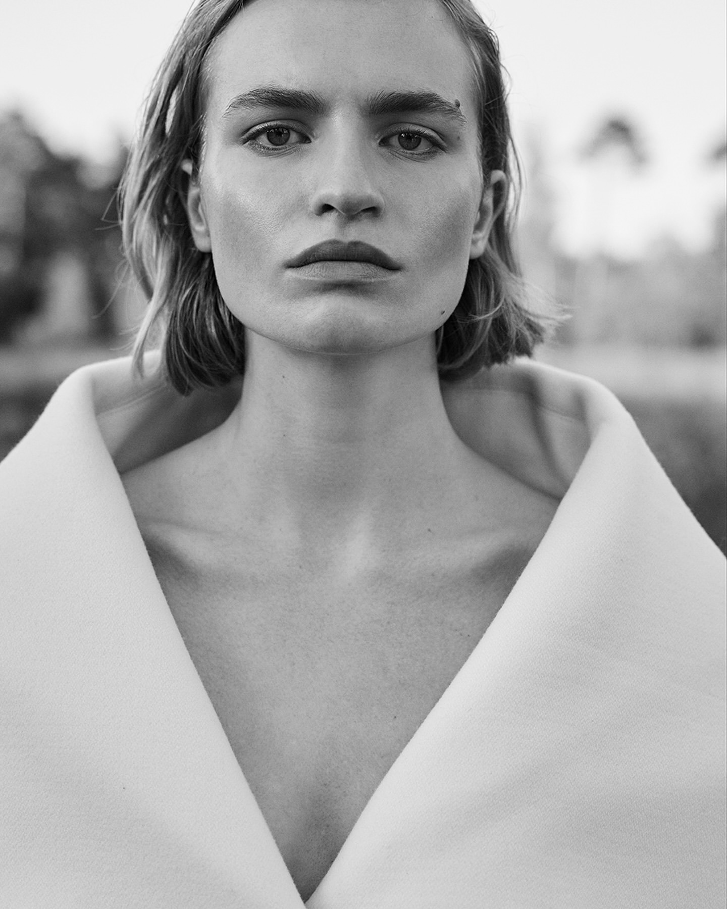 Striking black and white portrait of a young adult with short blonde hair and a white jacket against an out-of-focus outdoor background.