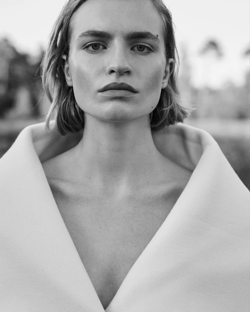 Striking black and white portrait of a young adult with short blonde hair and a white jacket against an out-of-focus outdoor background.