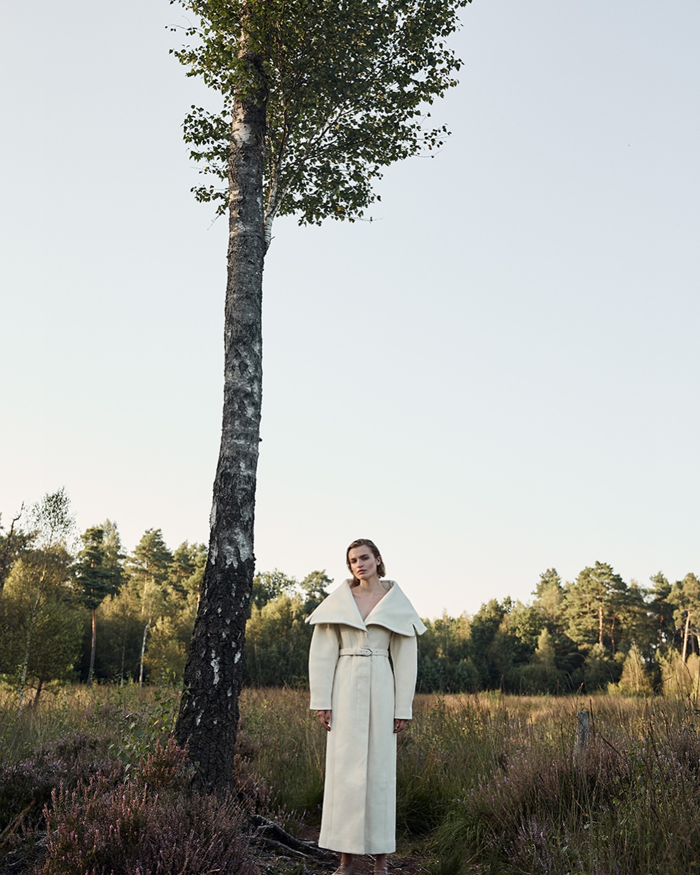 A model in an off-white coat stands near a tall birch tree in a field, with a distant line of trees beneath a light sky.