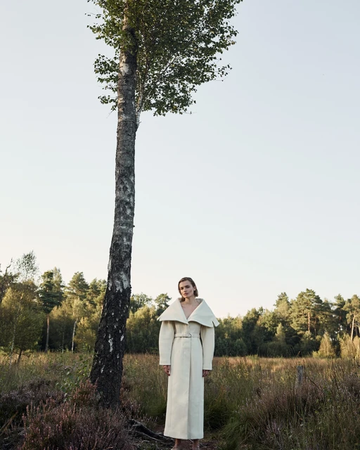 A model in an off-white coat stands near a tall birch tree in a field, with a distant line of trees beneath a light sky.