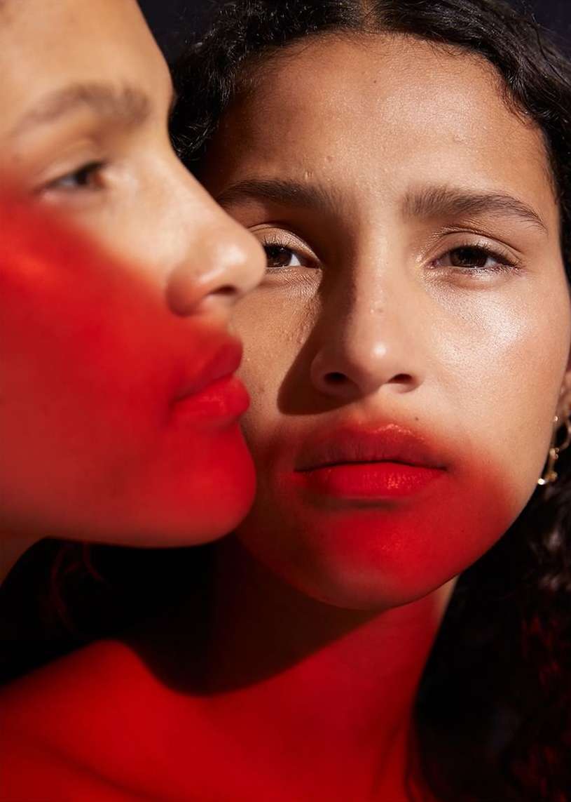 Close-up studio portrait featuring two young adult female models with curly hair and red lighting, set against a dark background.
