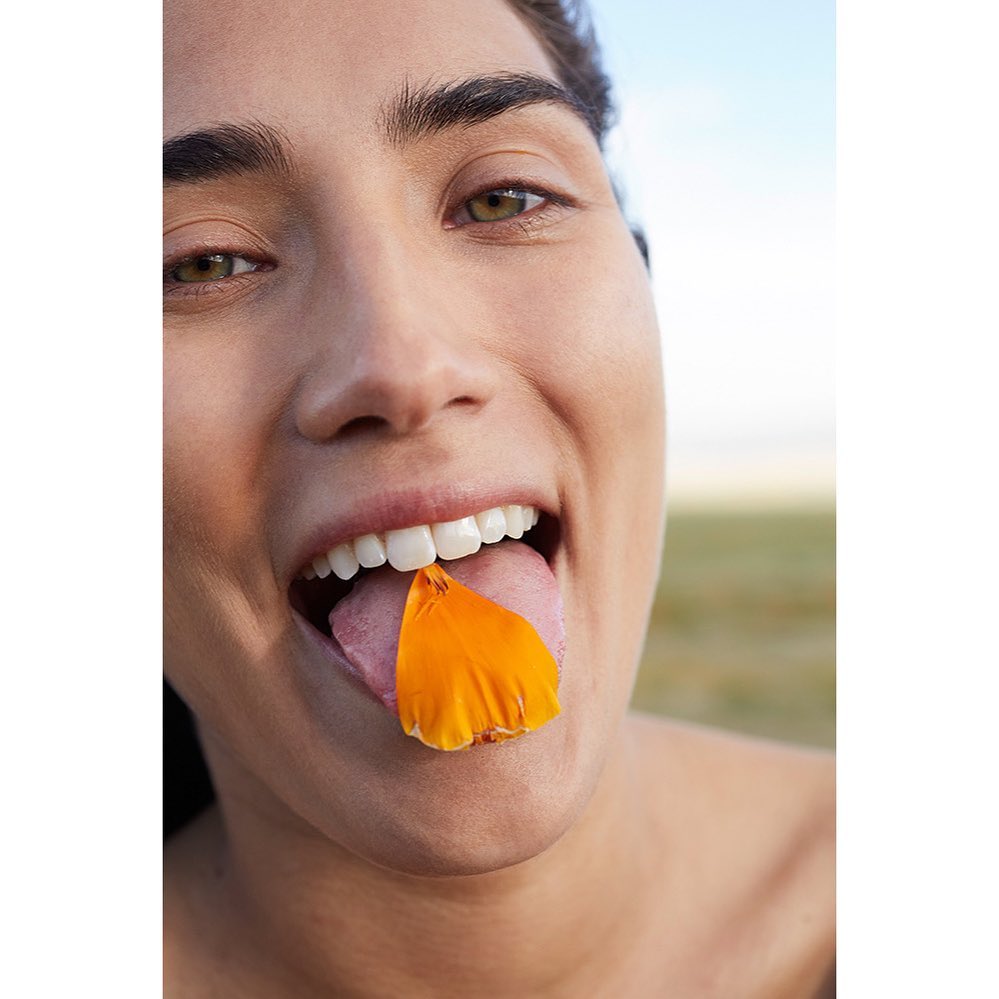 Close-up of a woman with a cheerful expression holding an orange petal on her tongue, outdoors in natural light with a plain background.