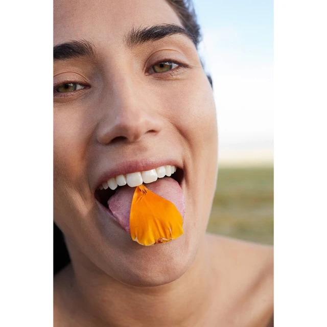 Close-up of a woman with a cheerful expression holding an orange petal on her tongue, outdoors in natural light with a plain background.
