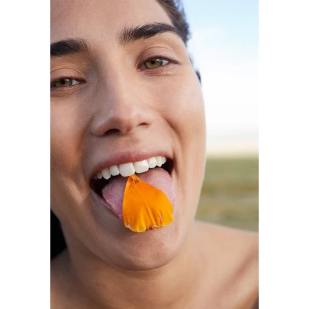 Close-up of a woman with a cheerful expression holding an orange petal on her tongue, outdoors in natural light with a plain background.