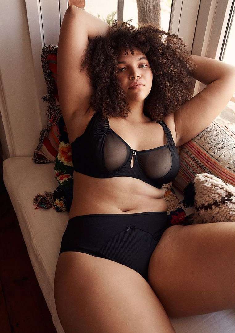 Adult female model with curly hair in black lingerie, reclining on a patterned bench indoors near a window with natural light.