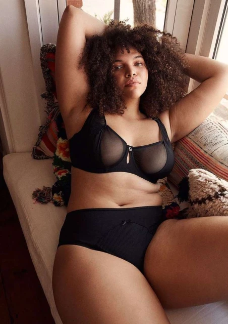 Adult female model with curly hair in black lingerie, reclining on a patterned bench indoors near a window with natural light.