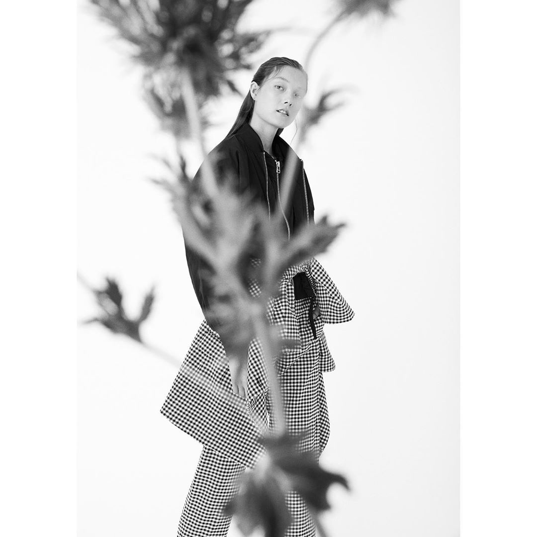 Fashion shot of a young adult female model in a black bomber jacket and gingham pants, posing against a white background with foliage.