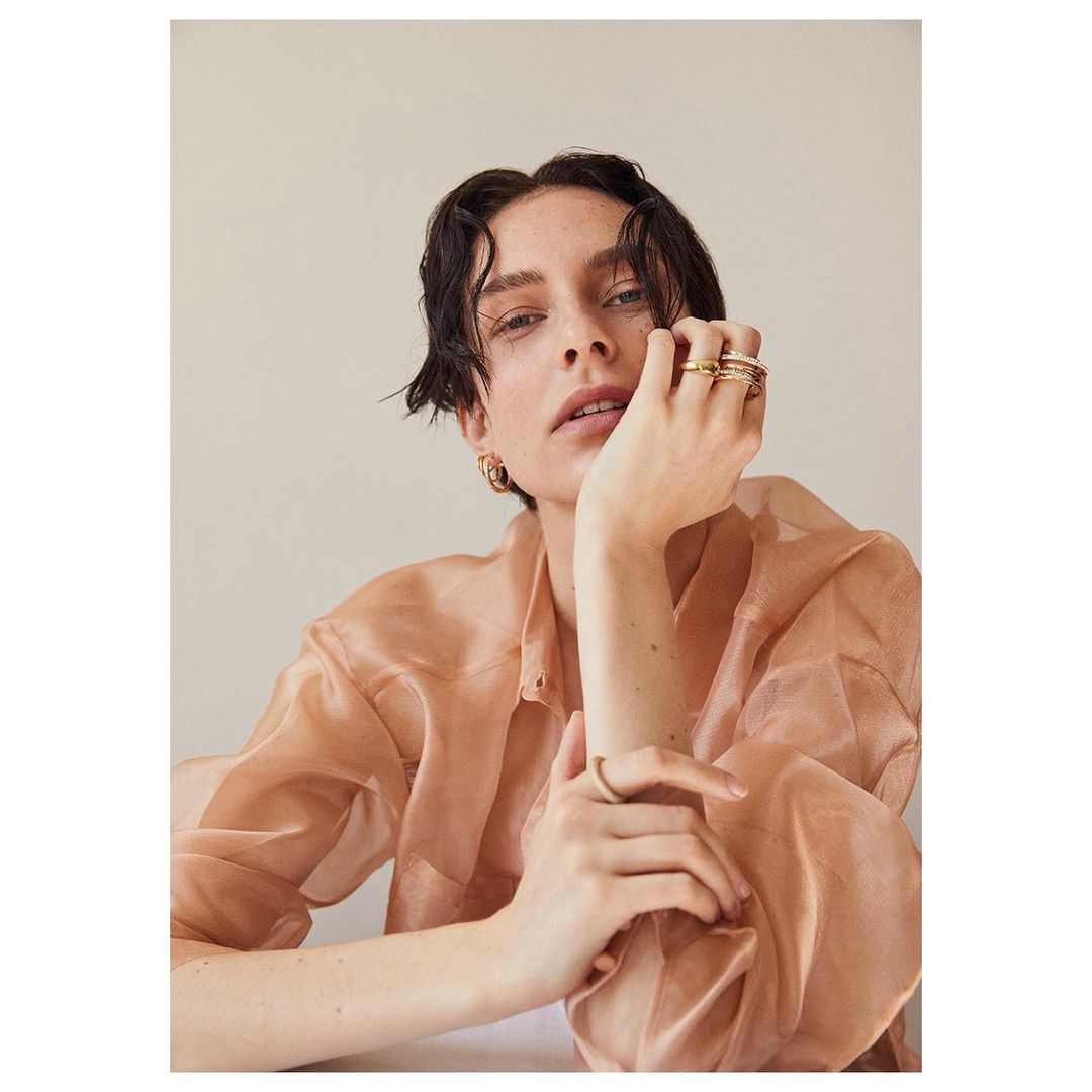 Portrait of an adult female in a peach-colored top with gold rings and earrings, posing indoors against a cream-colored backdrop, eye-level angle.