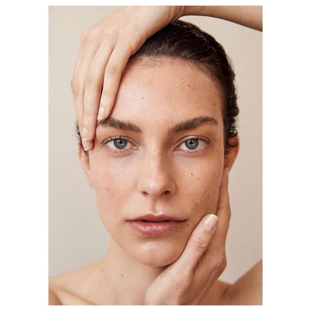 Close-up portrait of a young adult woman with freckles, blue eyes, and hands framing her face, against a neutral beige background.