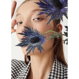 Close-up portrait of a woman wearing a black and white gingham blazer, partially obscured by blue thistle flowers.
