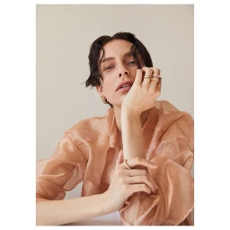 Portrait of a woman wearing a sheer peach blouse with stacked gold rings and hoop earrings against a neutral background.