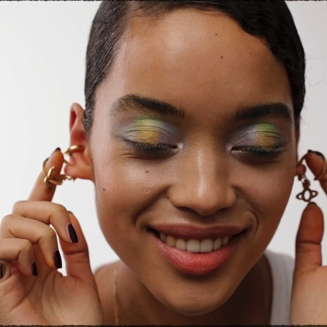Close-up portrait of a smiling woman with iridescent eyeshadow holding her earrings against a white background.