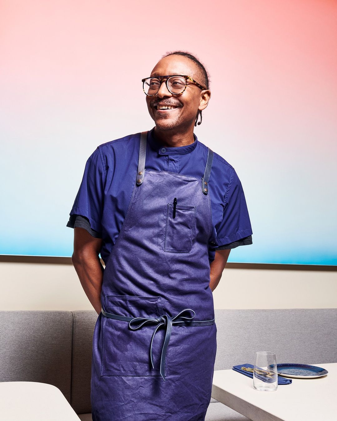 Smiling adult man wearing glasses and a blue apron stands in a brightly lit restaurant interior with a colorful background.