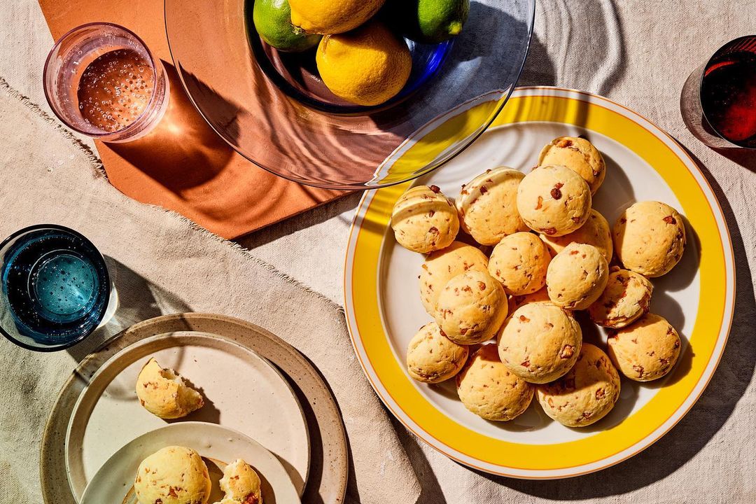 Overhead shot of a yellow plate with Pão de Queijo, lemons, limes, and glasses of water and wine on a textured beige tablecloth.