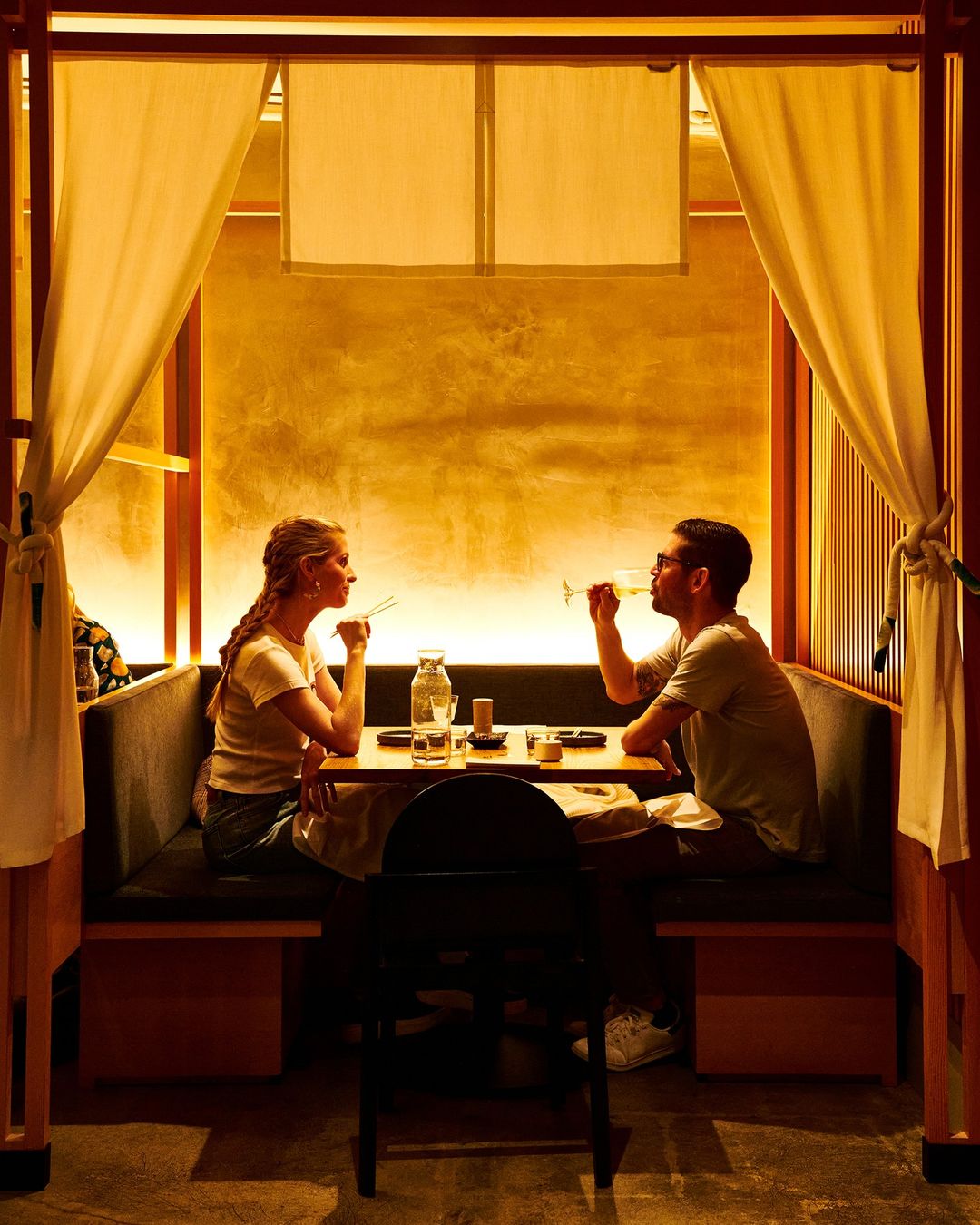 A couple dines at a restaurant booth with golden wallpaper, enjoying drinks in a warmly lit, intimate atmosphere with cream-colored curtains.