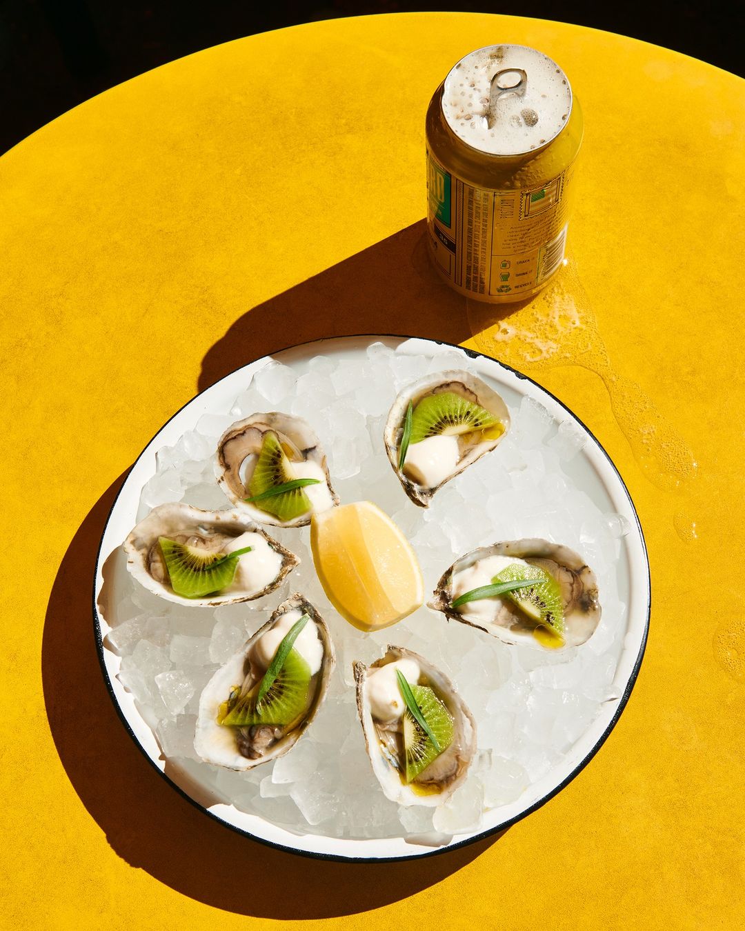Overhead shot of oysters on crushed ice with kiwi and lemon, presented on a round white plate atop a bright yellow table with a can.