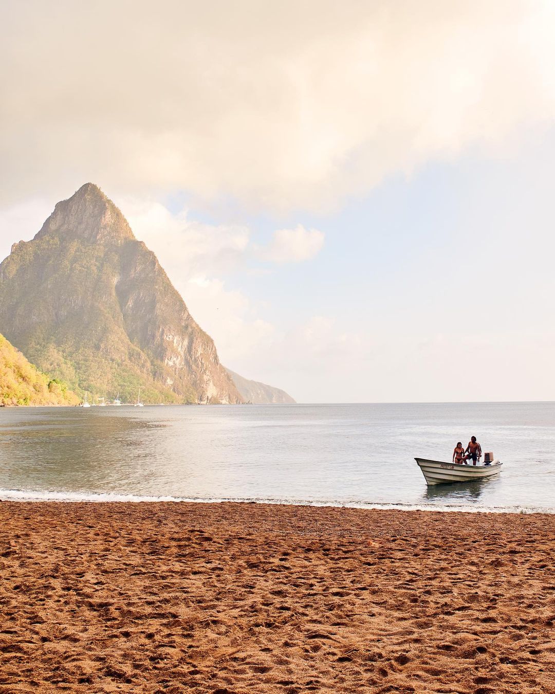 Two adults and a child in a small boat near the beach, with a large mountain in the background on a partly cloudy day.