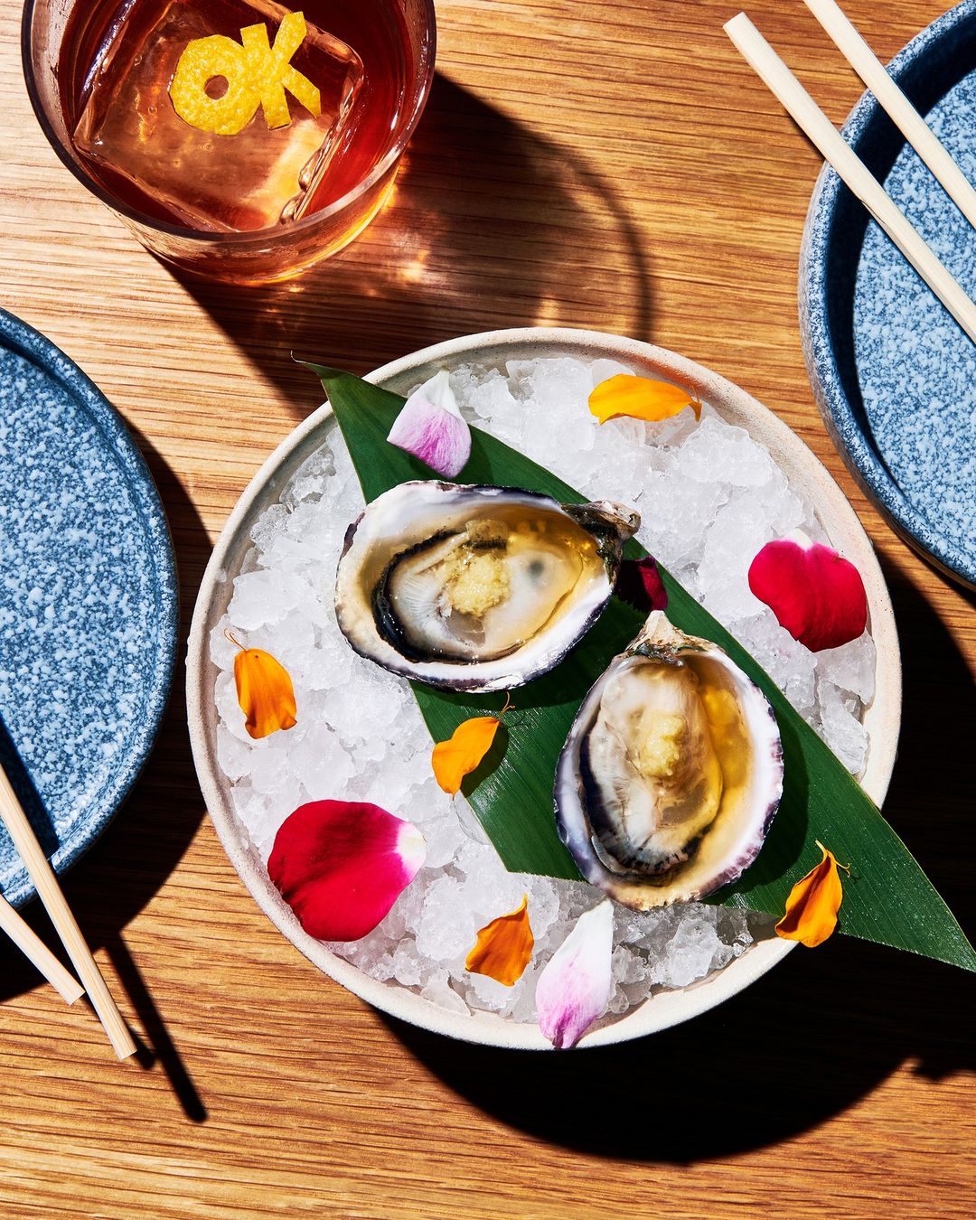 Overhead shot of two oysters on ice with floral petals on a plate, accompanied by a cocktail and chopsticks on a wooden table.