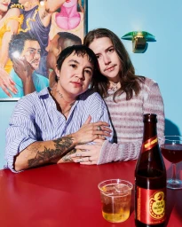 Two people seated closely together at a red table with a beer bottle and glass, against a blue wall with a painting.