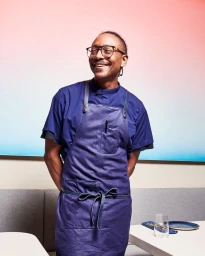 Smiling Black man wearing a blue chef apron and tortoiseshell eyeglasses against a pink and white gradient backdrop.