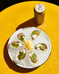 Oysters on crushed ice garnished with kiwi and a lemon wedge beside a drink can on a bright yellow table.