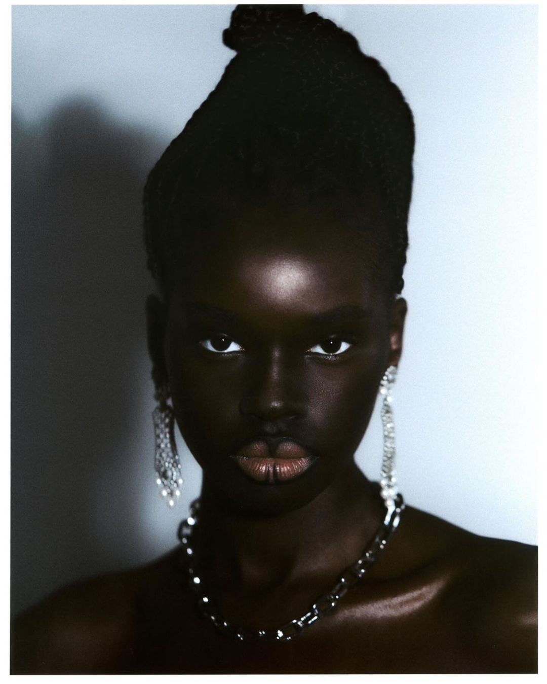 Close-up portrait of a Black woman with elaborate hairstyle, dangling earrings, and necklace against a soft gray background. Beauty shot.