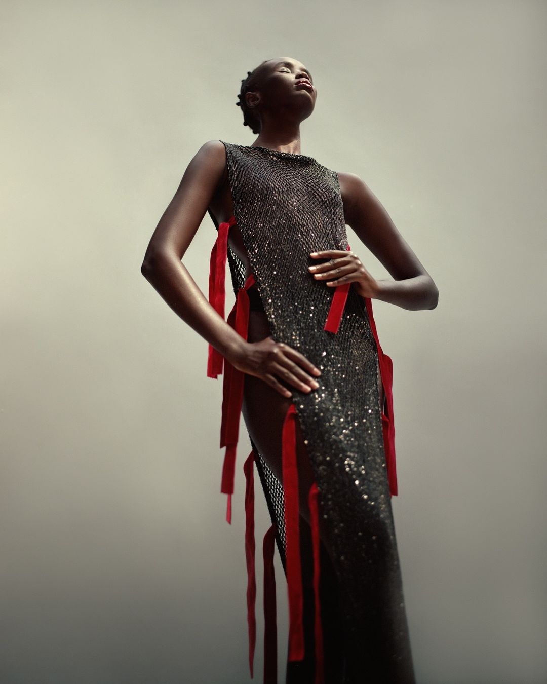 Elegant, dark-skinned woman in long, sparkling gown with red ribbons, head tilted back in a confident pose, studio lighting, high angle.