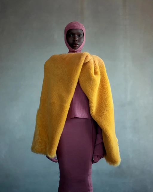 Dark-skinned adult female in a pink ribbed fitted dress and head covering poses with a yellow fur wrap against a gray backdrop.