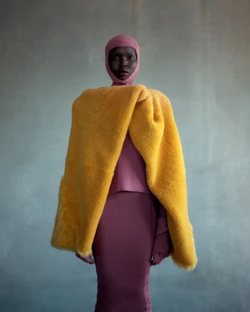 Dark-skinned adult female in a pink ribbed fitted dress and head covering poses with a yellow fur wrap against a gray backdrop.