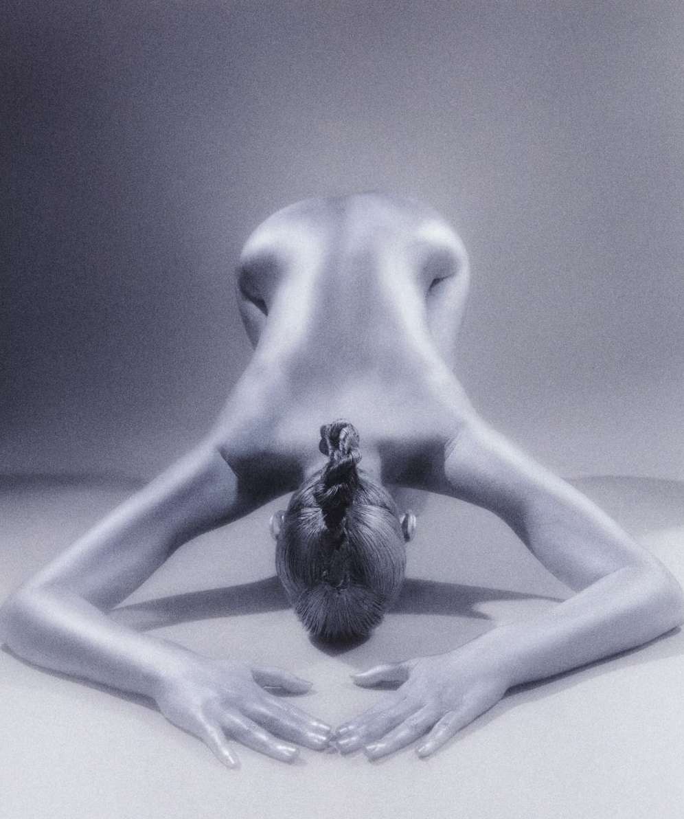 Back view of bare-backed adult female bending forward with interlocked fingers resting on a grey surface in a neutral environment.