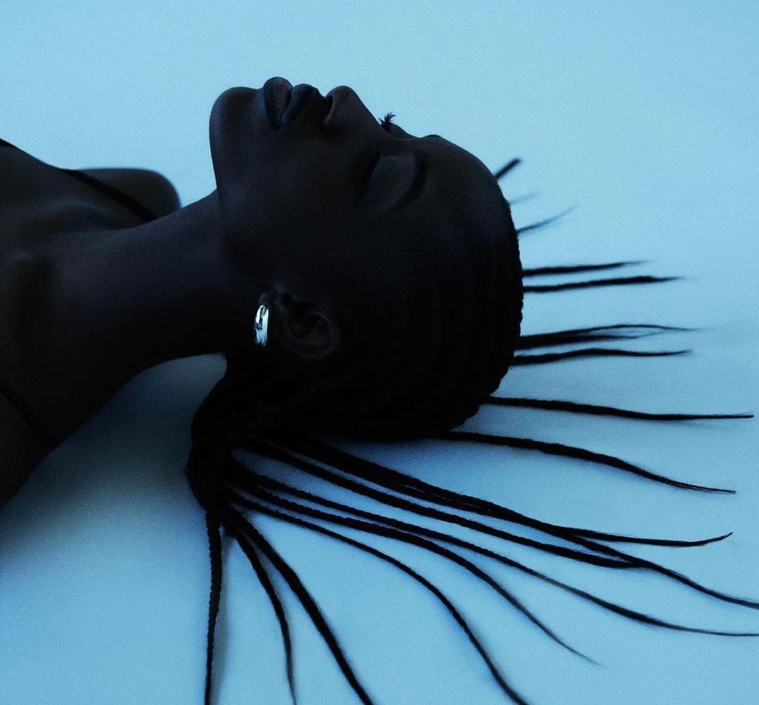 Monochromatic portrait of a young African-descent woman with braids, laying against a light blue surface with a serene facial expression.