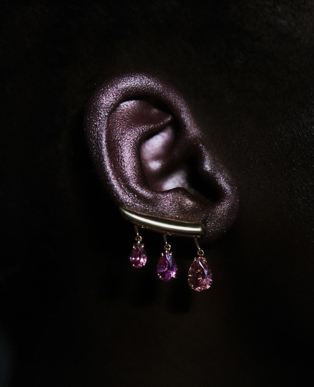 Close-up shot of a human ear adorned with dangling pink gemstone earrings, set against a deep black background, showcasing beauty and jewelry.