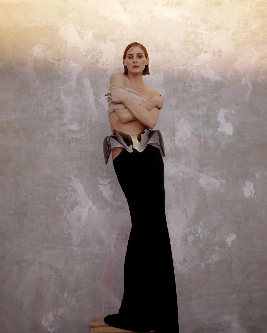 Woman in a black maxi skirt and metallic peplum corset standing against a mottled textured wall.