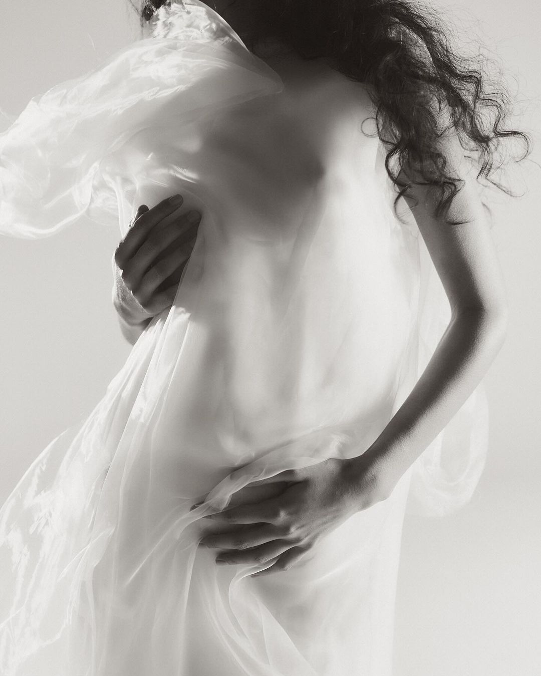 Model draped in sheer white fabric with long curly hair captured in a moody monochrome portrait by Myrthe Giesbers.