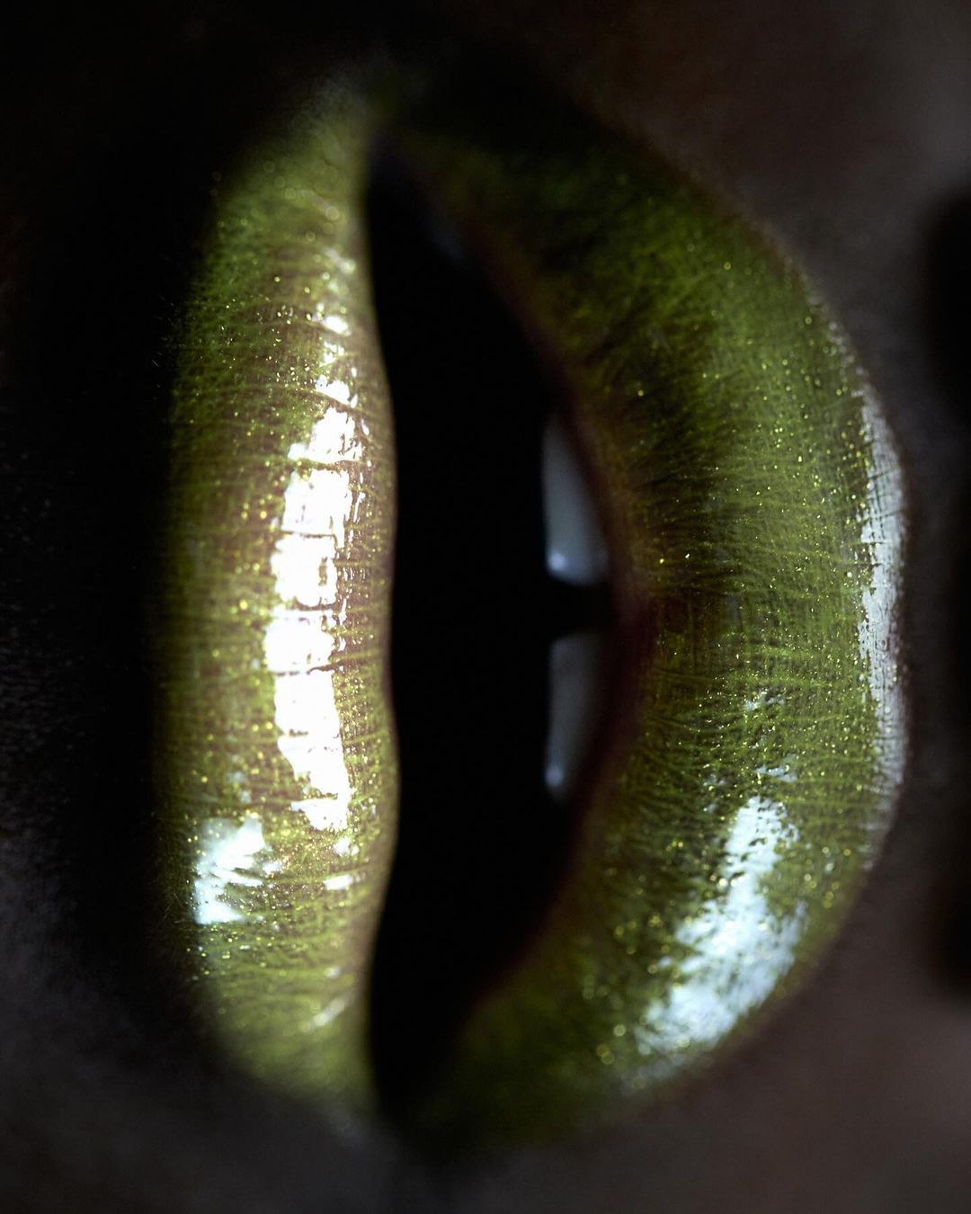 Extreme close-up of human lips covered in shimmering, glossy, textured gold lipstick and partially visible teeth in dark environment.