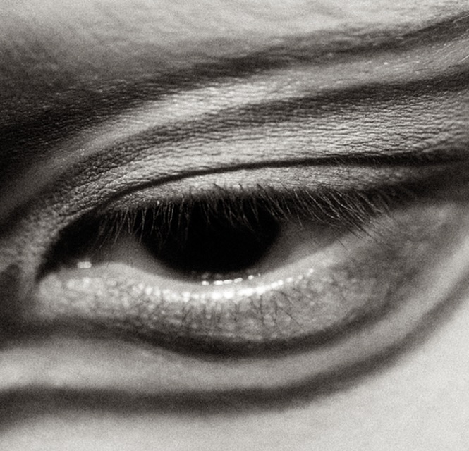 Close-up black and white photograph of a human eye with visible eyelashes, eyebrow and skin texture, showcasing a natural expression.